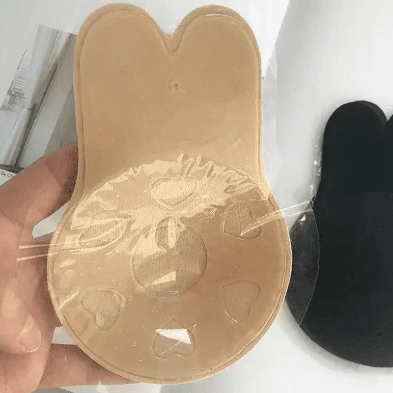 Sutiã adesivo de silicone bege com textura de corações em detalhe, sendo segurado contra fundo branco.
