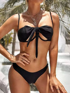  mulher vestindo um biquíni preto com um laço na parte da frente. Ela usa várias joias, incluindo um colar com pingentes. Ao fundo, há vegetação típica de ambiente tropical.