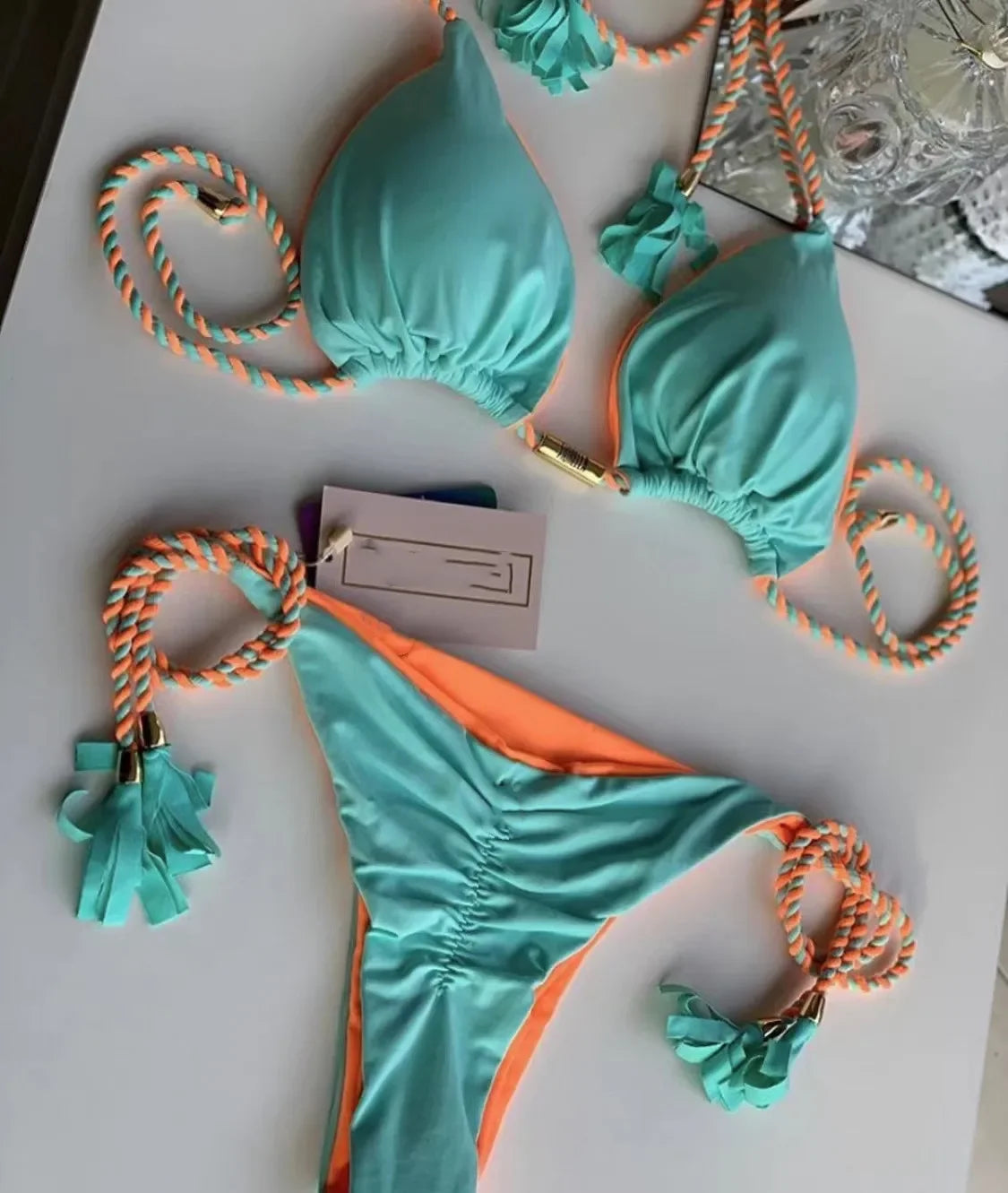 Bikini em tons de verde e laranja com detalhes decorativos em cordas trançadas. Inclui uma etiqueta de preço, sugerindo que se trata de um produto à venda.