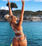 Mulher em biquíni colorido estampado fazendo pose em barco, mar azul e costão rochoso ao fundo