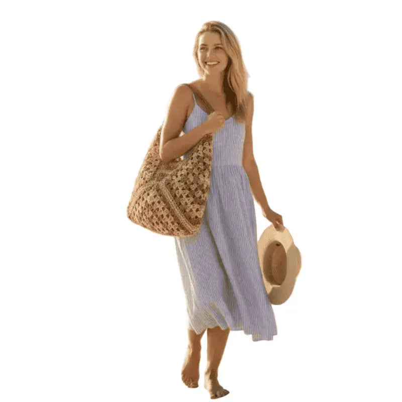 Mulher usando vestido listrado azul e branco com bolsa de crochê bege e chapéu na praia
