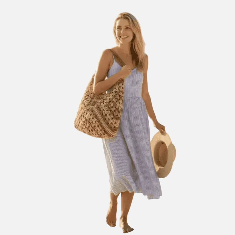 Mulher usando vestido listrado azul e branco com bolsa de crochê bege e chapéu na praia