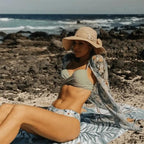 Conjunto praia: biquíni verde menta com saída de praia  floral, chapéu de palha decorado, em cenário praiano com rochas e mar