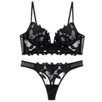 Conjunto de lingerie preto com renda floral transparente, sutiã meia taça e calcinha fio dental com acabamento ondulado.