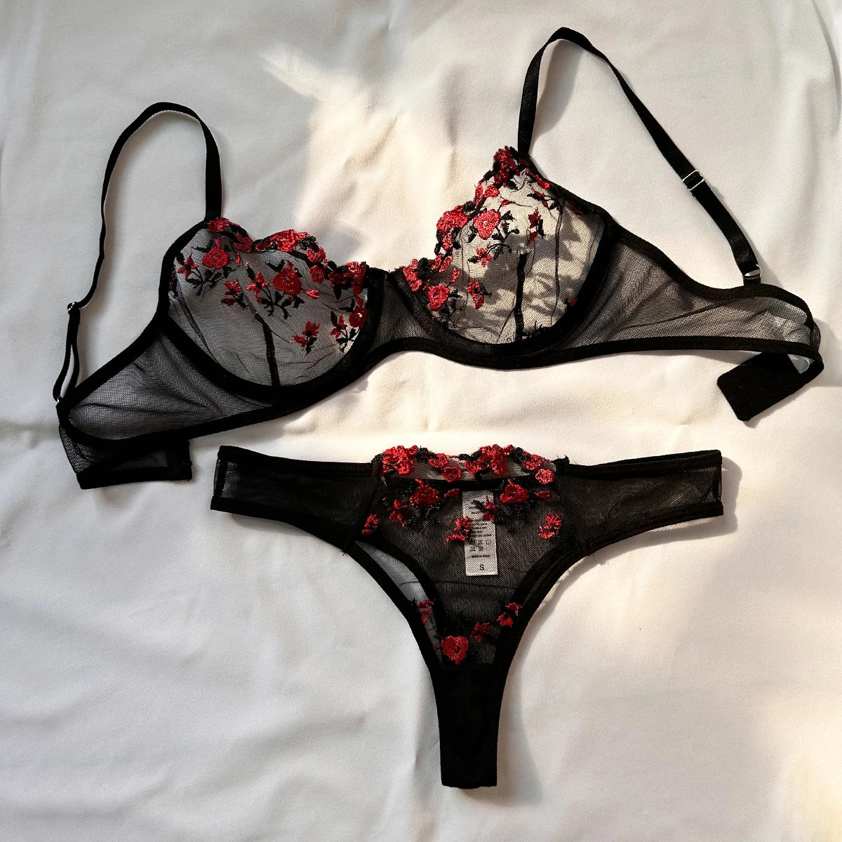 O conjunto de lingerie é composto por um sutiã e uma calcinha com uma elaborada estampa floral vermelha em um tecido semi-transparente preto