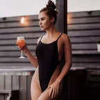 Mulher jovem usando maiô preto de alças finas segurando taça com drink laranja em ambiente moderno.