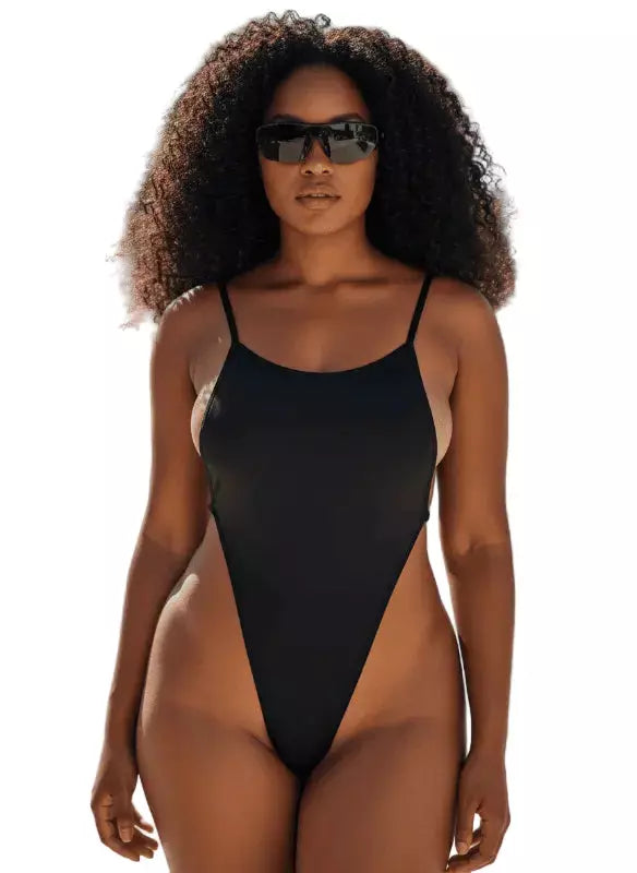 Uma mulher com cabelos cacheados usando óculos escuros e maiô preto de alças finas em fundo claro.