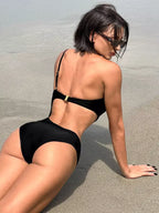 Maiô preto com recorte nas costas e fivela dourada, mulher sentada na areia da praia, estilo sofisticado.