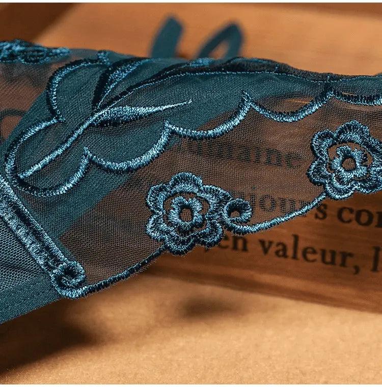 Detalhe de calcinha em tule azul petróleo com bordado floral metálico sobre fundo marrom.